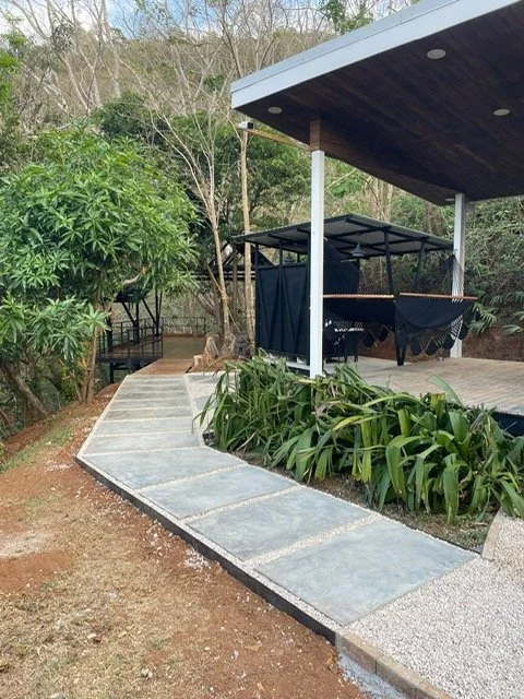 A covered outdoor patio area with a swing, surrounded by greenery and garden plants.