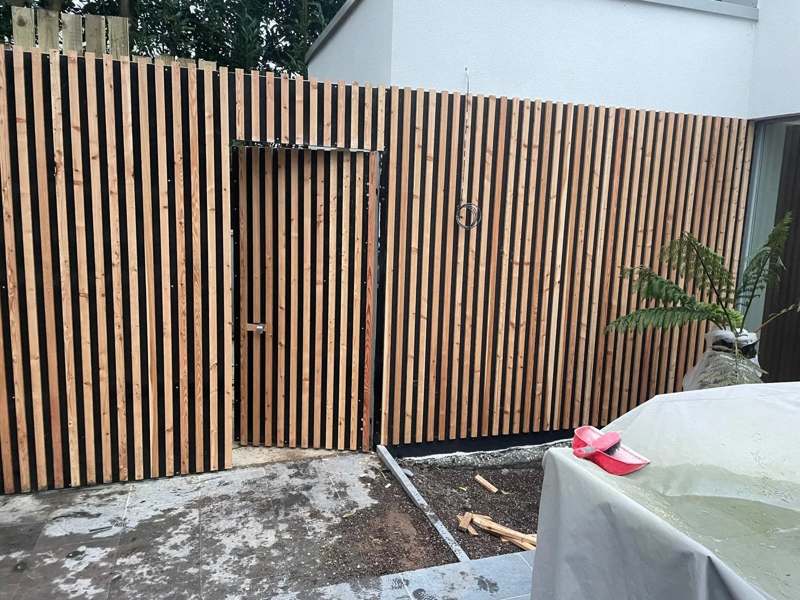 A wooden slat privacy fence in a backyard with a partially installed gate, construction materials and a covered table with pink slippers on top.