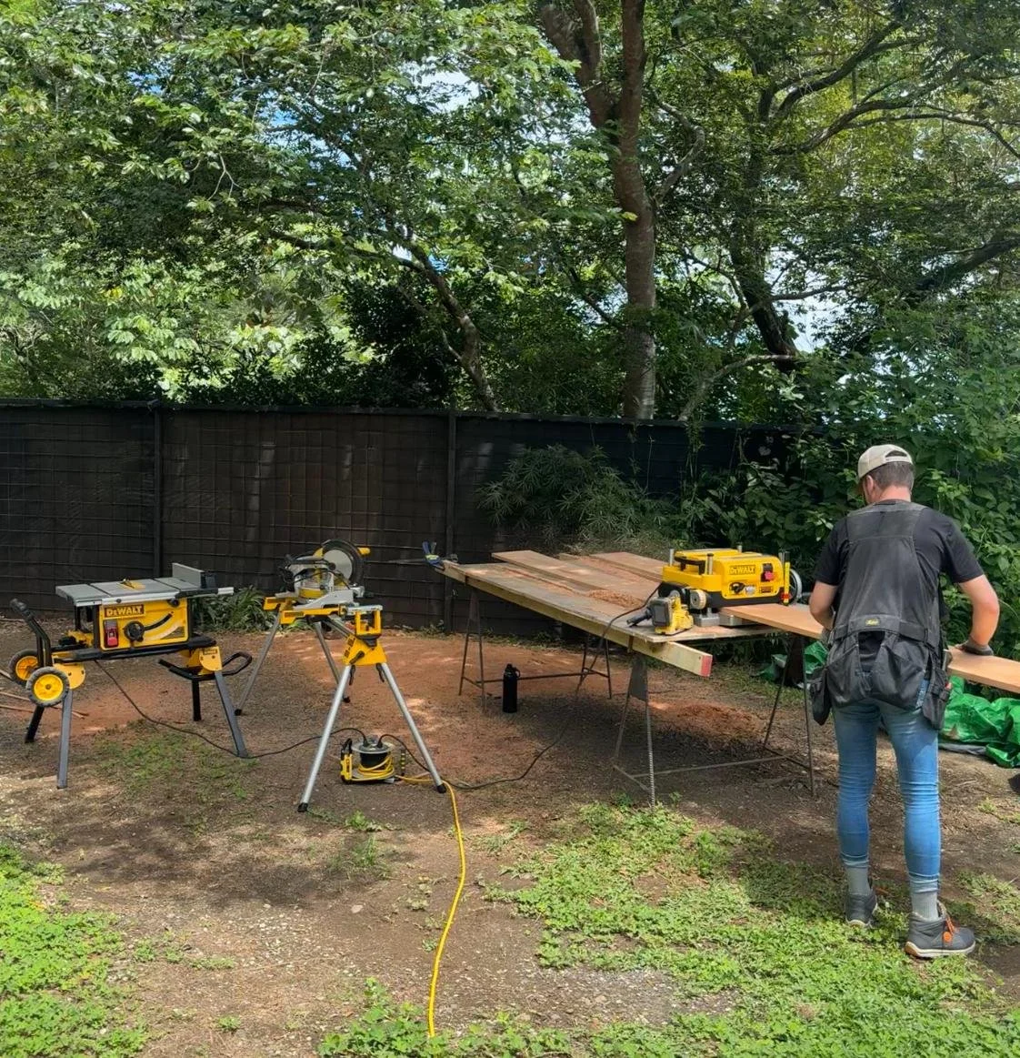 Person working on wood project with power saws and other tools in outdoor backyard with trees and black privacy fence.