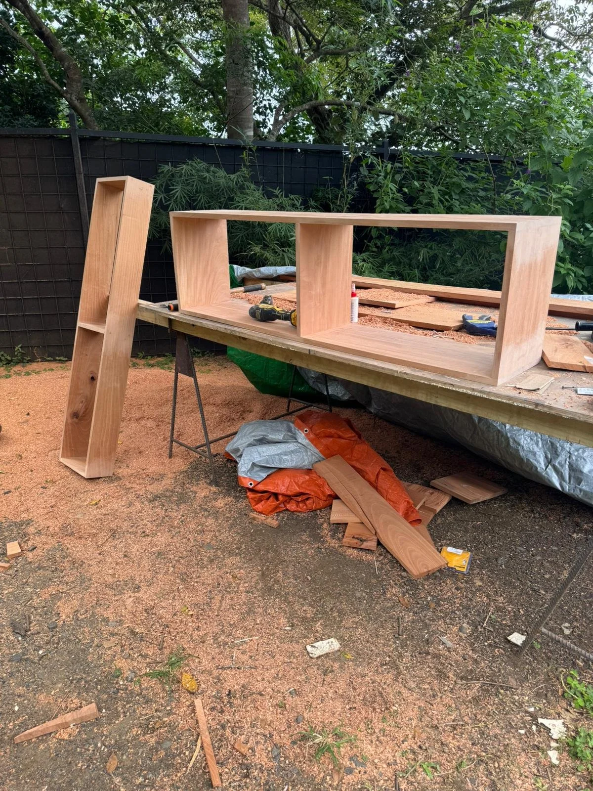 Wooden furniture being constructed outdoors with tools and supplies on a worktable. Parts of the furniture and sawdust on the ground.