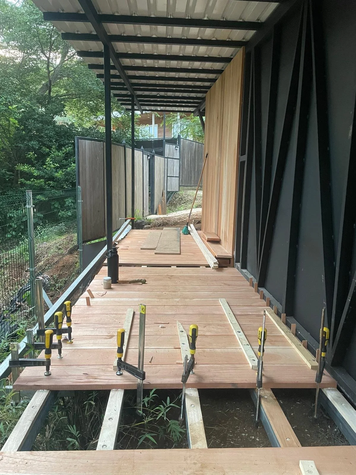 Under construction of a wooden deck or porch, with partially assembled wooden boards and clamps, in an outdoor residential area next to a house with black wall and wooden siding, enclosed by wooden and metal fencing, surrounded by greenery.