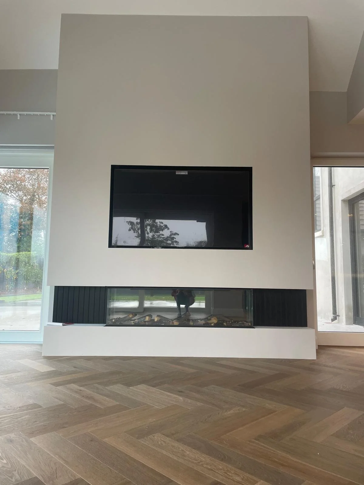 Modern fireplace with a black glass screen, located below an off-white wall-mounted television, in a room with wooden herringbone-patterned floors and large windows.