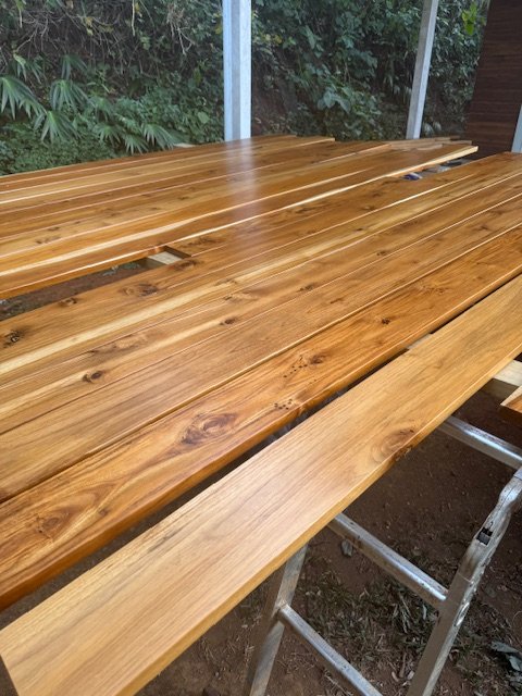 Several wooden planks are laid on a work table, with an outdoor background of trees and bushes visible through a screened enclosure.