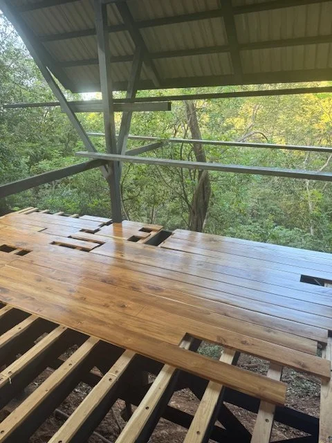 Wooden deck or floor in progress with some planks installed and others waiting to be put down, overlooking a wooded area with trees, under a metal roof structure.