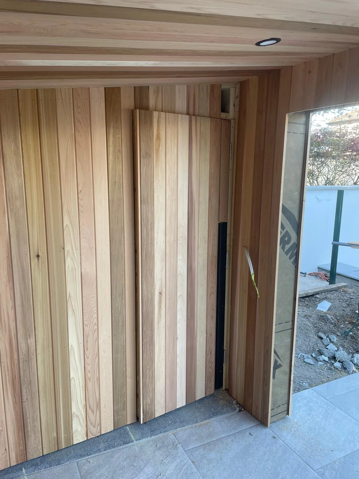 Interior of a building under construction with wooden walls and a partially installed door, with a window on the right side showing an outdoor area.