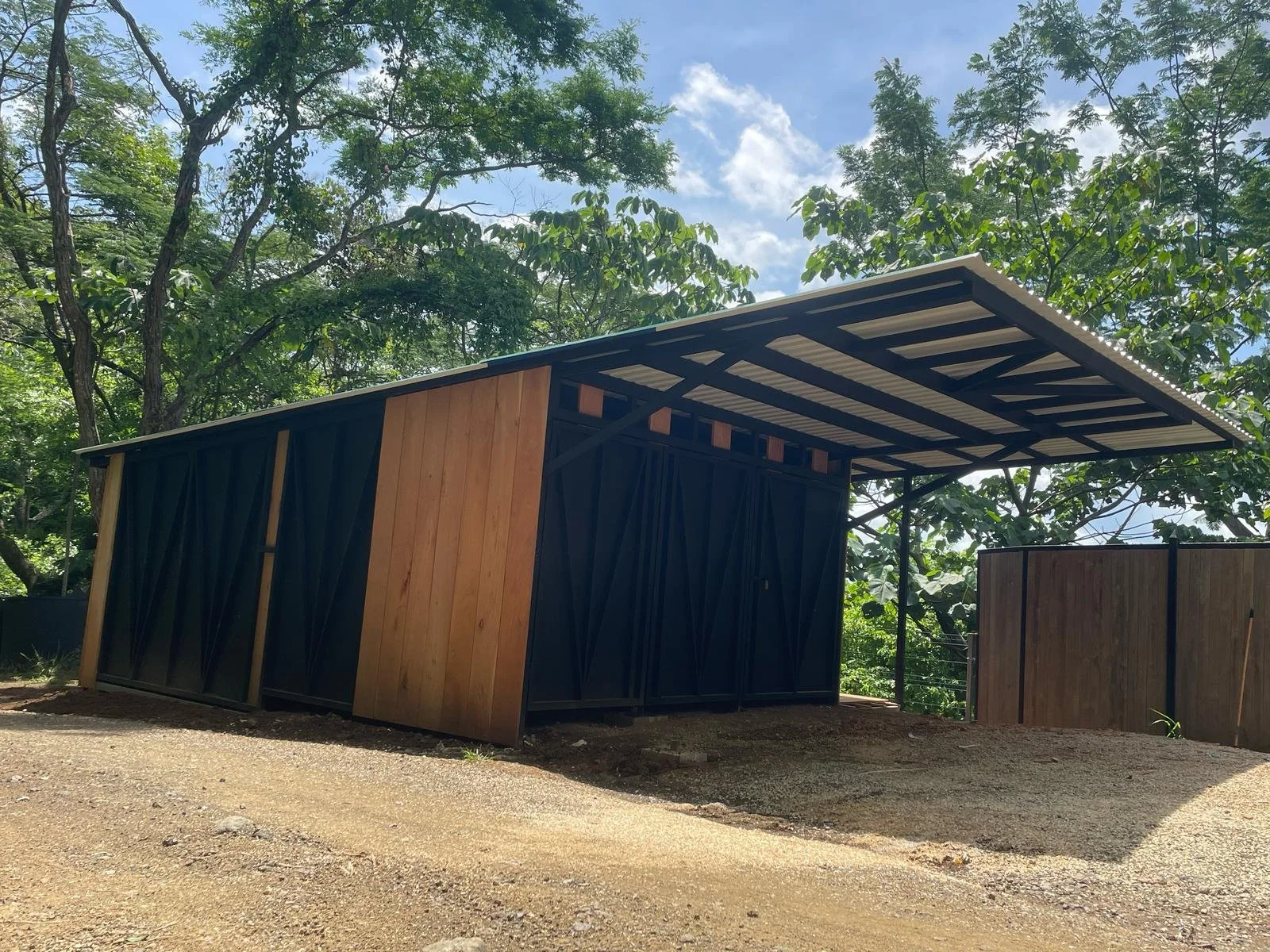 A small modern shed with a black metal exterior and wooden panels, set in a wooded area with gravel ground and a partly cloudy sky overhead.