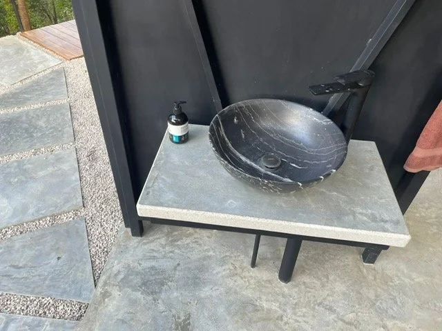 Outdoor sink with a black vessel basin, a soap dispenser, and a black faucet, against a black privacy screen with a concrete countertop and stone paving.