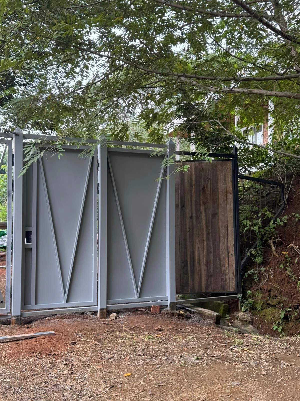 A partially constructed outside gate with metal and wooden sections, on uneven dirt ground, underneath leafy tree branches.