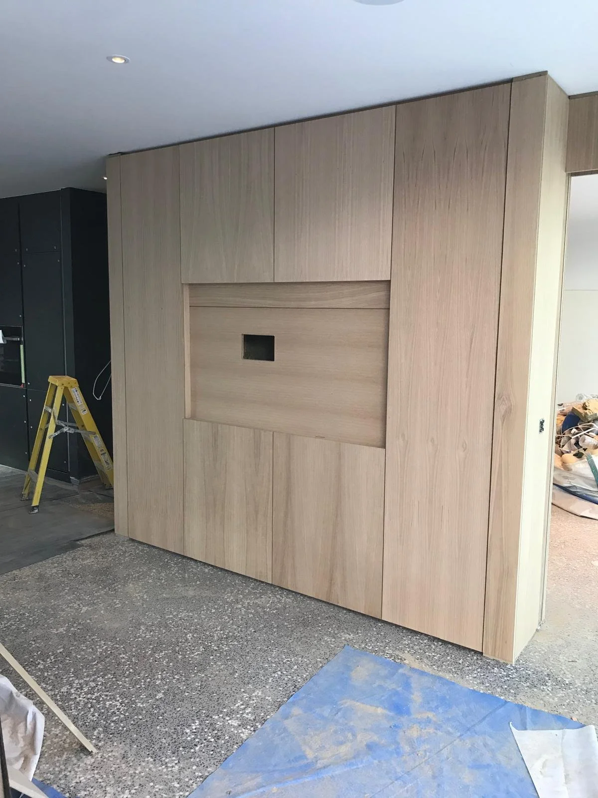 Interior space under construction with wood paneling on the wall and a small square cutout in the center, a ladder, and construction materials on the floor.