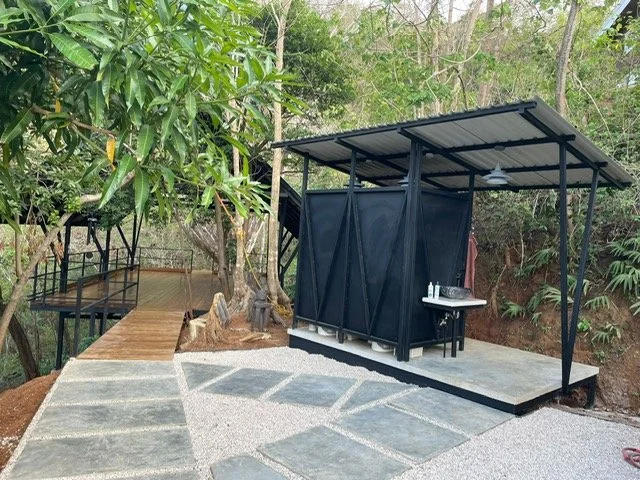 Outdoor shower under a metal roof with black curtains, next to a wooden deck and surrounded by trees.