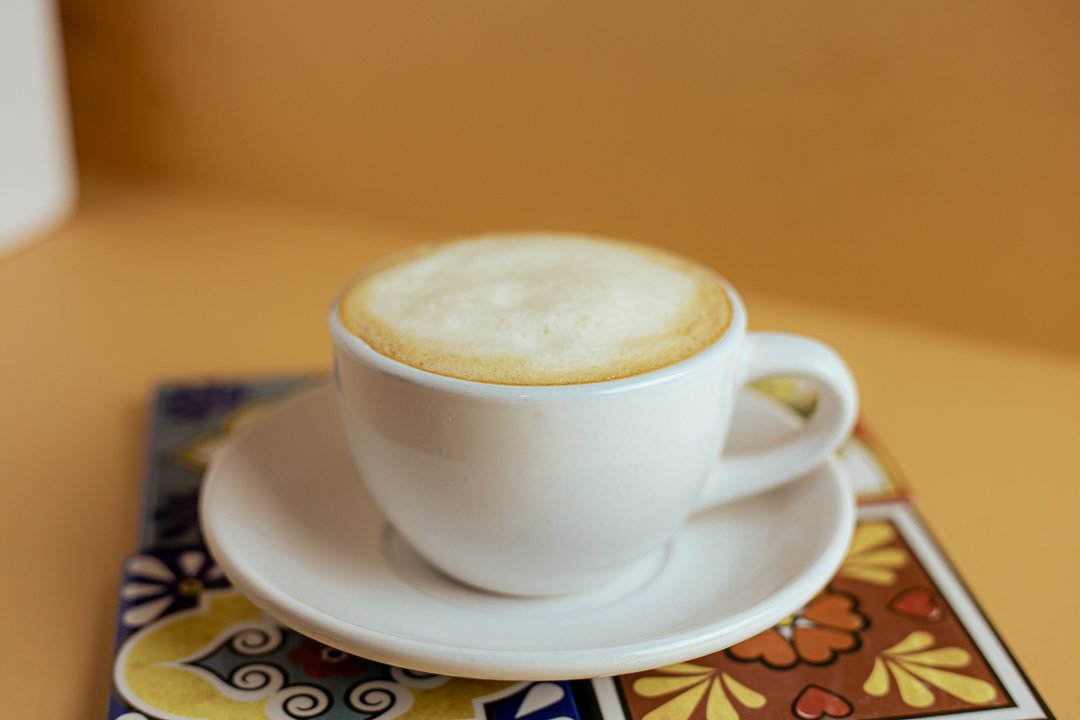 A white coffee cup filled with a frothy cafe con leche, sitting on a matching saucer on a colorful table with floral and geometric patterns. Louisville's cuban cafe and creamy cuban coffee.