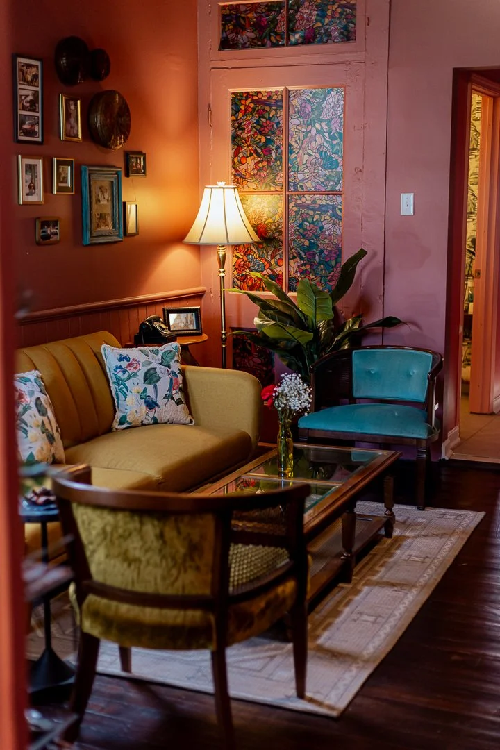 Cozy living room with a yellow sofa and floral pillows, blue armchair, glass coffee table with a flower vase, large potted plant, wall-mounted picture frames, a lit floor lamp, and stained glass window behind the plant.