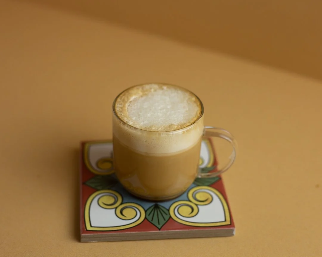 A clear glass mug filled with coffee topped with foam, sitting on a colorful decorative coaster on a yellow surface.