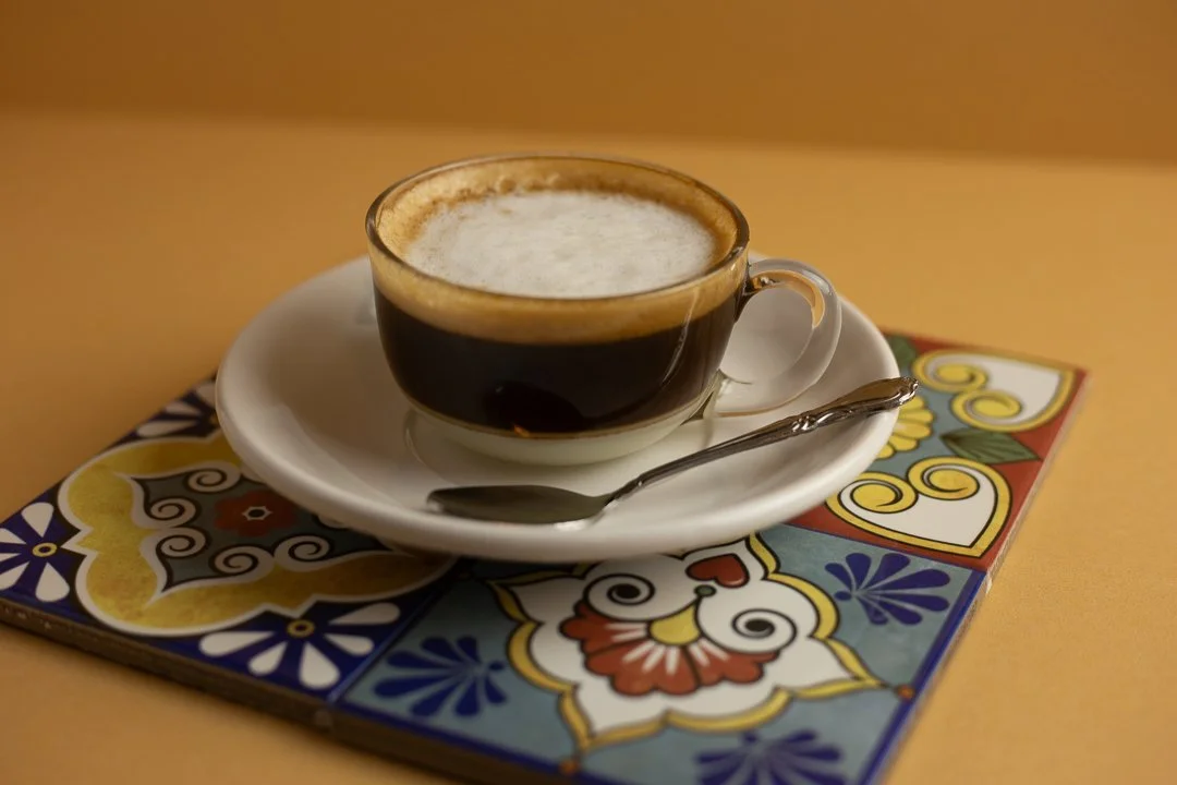 A cup of cuban cafecito with foam on top, served on a white saucer with a metal spoon, placed on a colorful decorative tile or coaster on a yellow surface. Cafe bombon in louisville, KY
