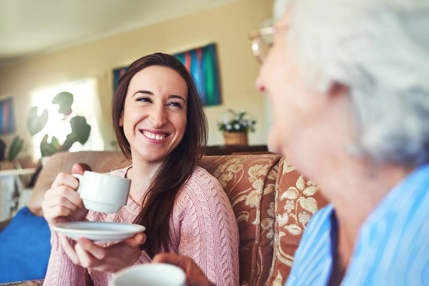 Young woman smiling while holding a cup and saucer, talking to an elderly woman who is partially visible.