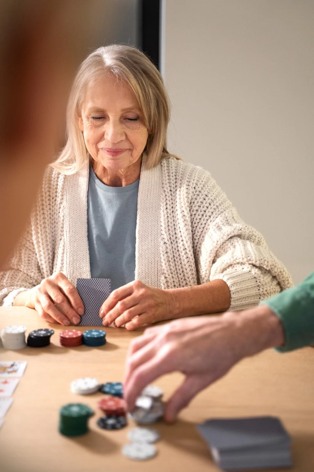 An elderly woman playing poker, smiling softly, with poker chips and playing cards on the table.