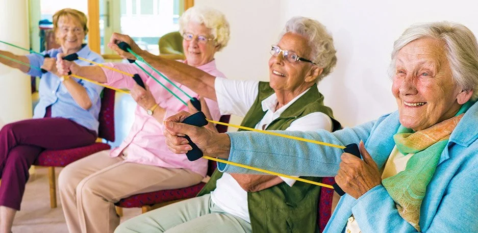 Four elderly women sitting in chairs doing resistance training with exercise bands, smiling and enjoying activity.