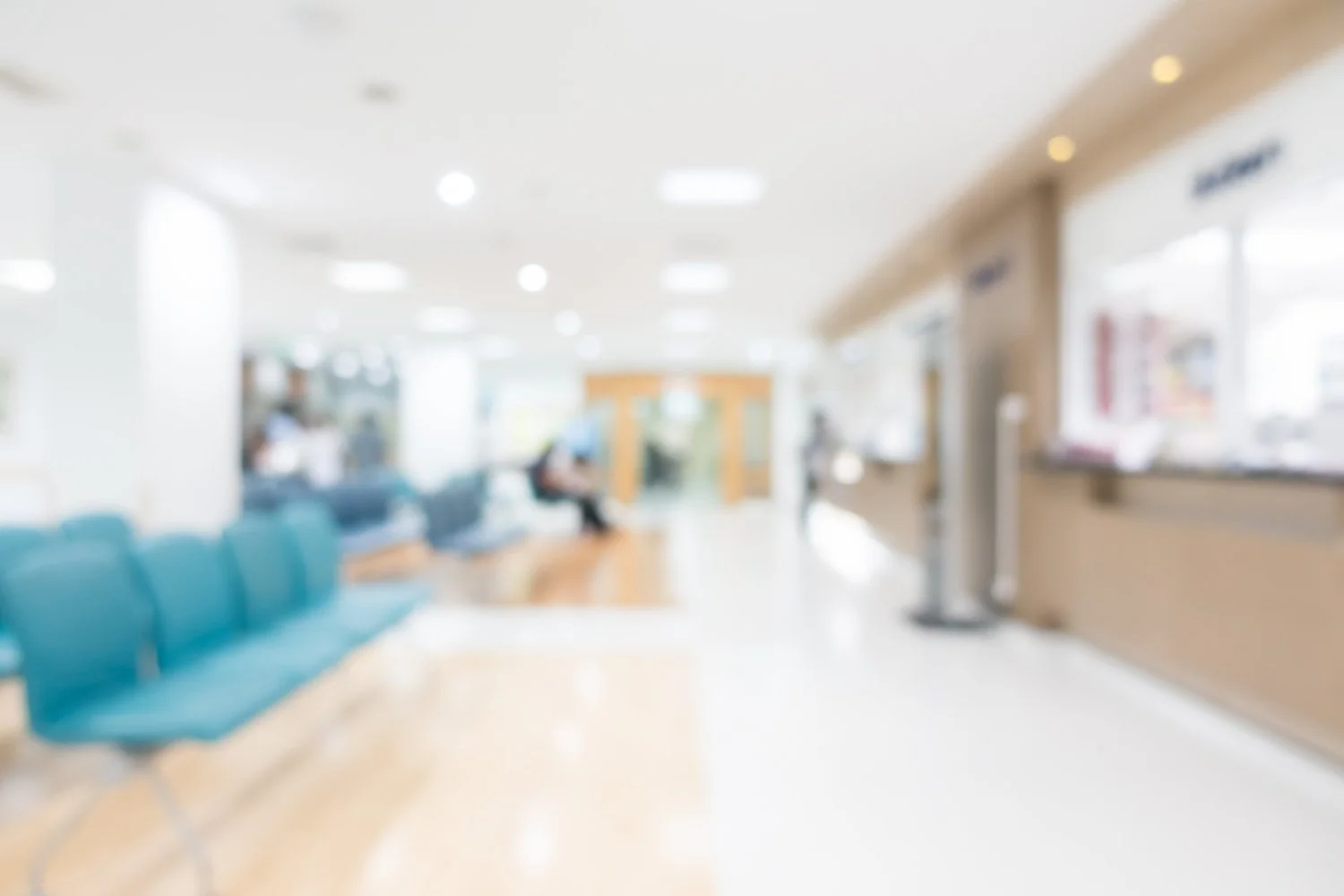 A blurry image of a hospital waiting room with blue chairs, a person sitting, and reception counter on the right.