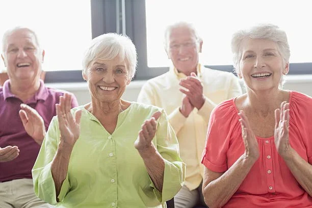 Four elderly people, two women and two men, smiling and clapping, sitting in a well-lit room.