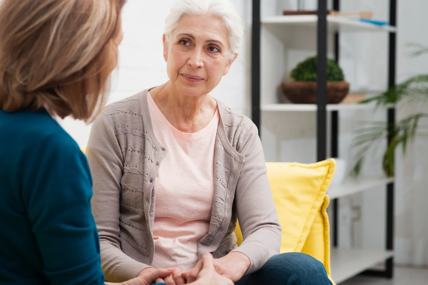 An elderly woman engaging in a heartfelt conversation with a caregiver or therapist in a bright, cozy room with shelves and plants in the background.