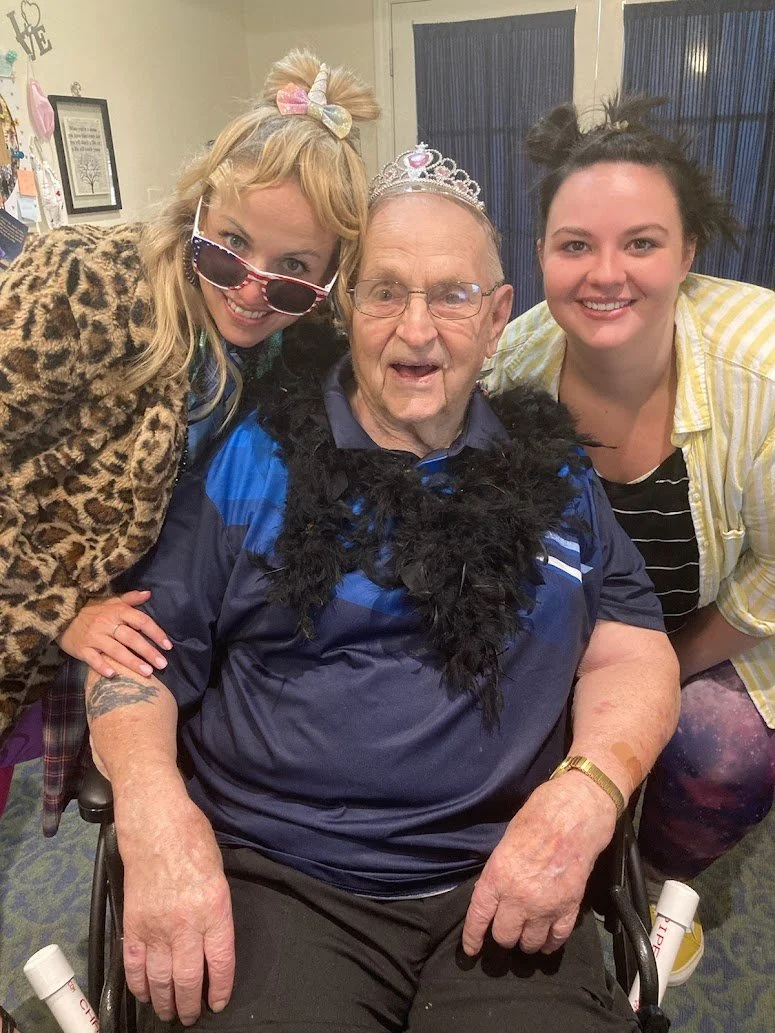 Three people celebrating a birthday or special occasion; an elderly man in a wheelchair wearing a tiara and a feather boa, flanked by two women smiling and leaning in close.