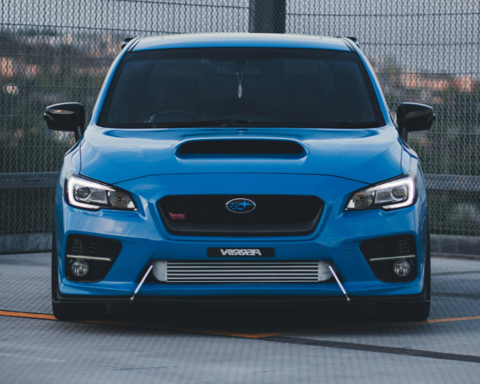 Front view of a blue Subaru WRX STI with hood scoop, modified front bumper, and intercooler visible through the grille, parked on a rooftop with a safety fence in the background.