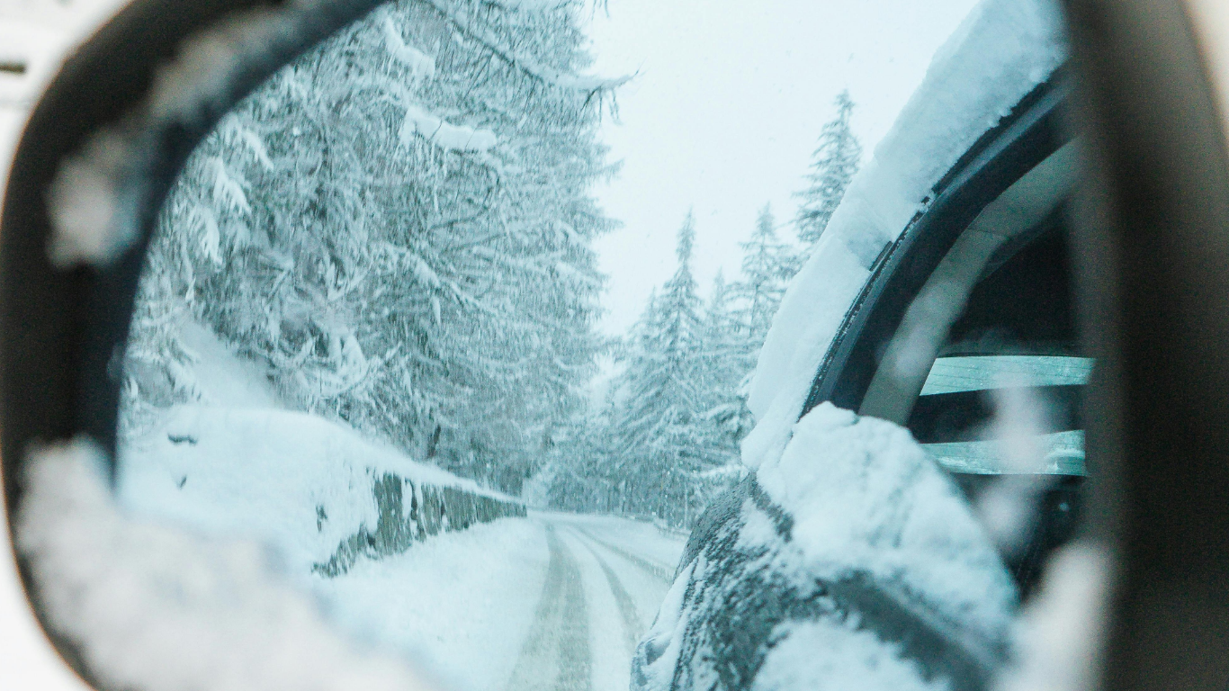Rearview mirror reflecting a snowy road with snow-covered trees and a snow-covered fence on the side.