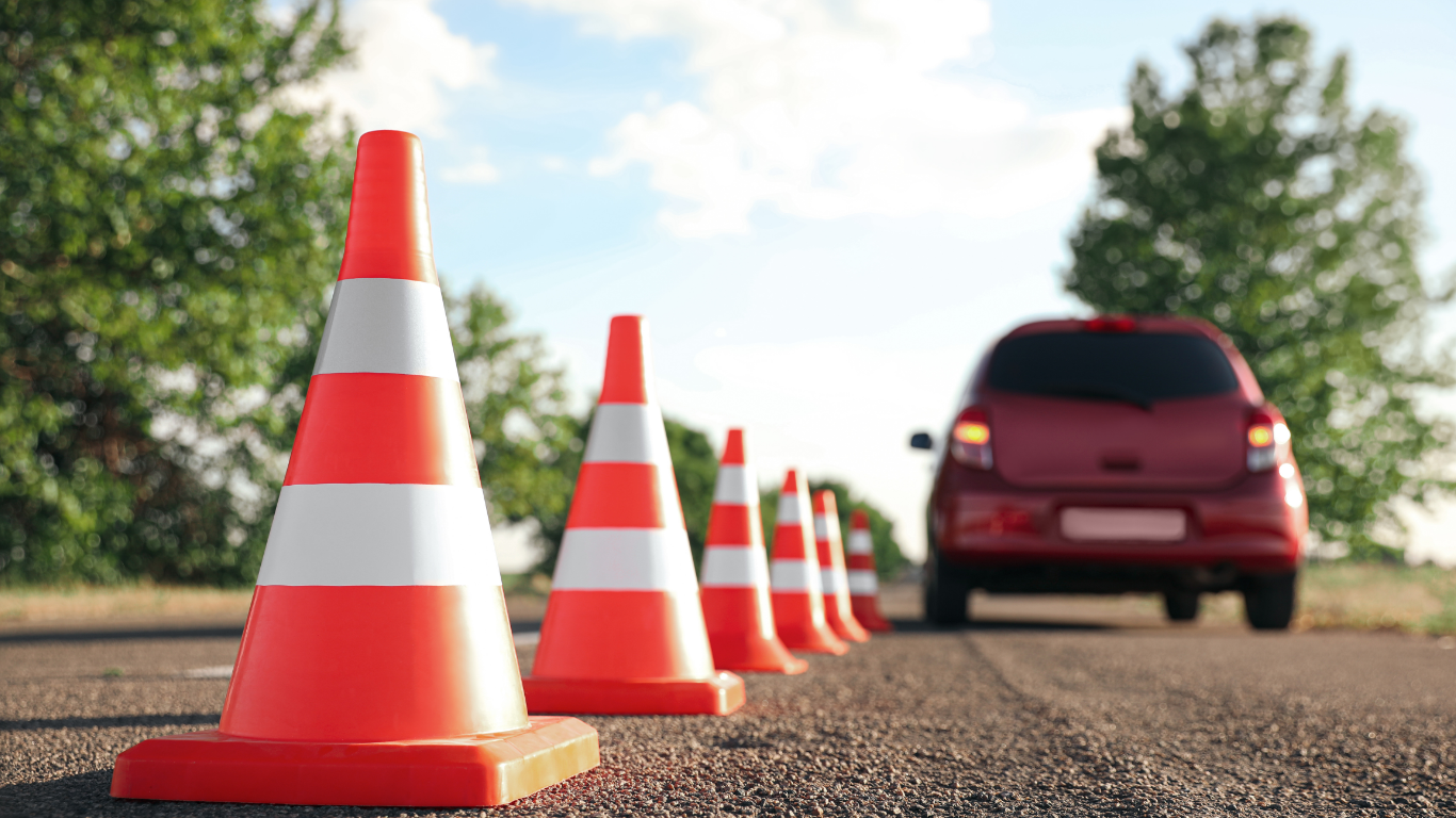 A line of orange safety cones on an asphalt road with a red car in the background, surrounded by trees and a partly cloudy sky.