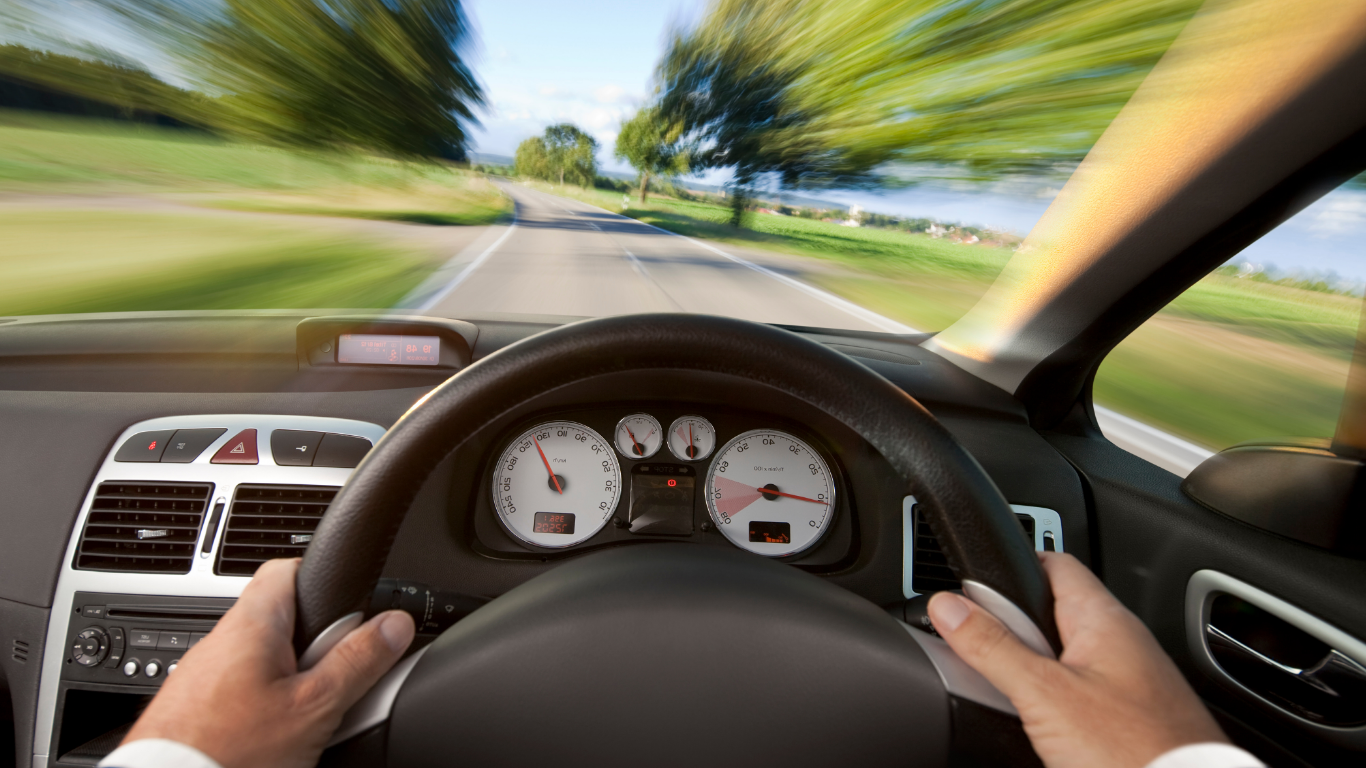 Driving on a rural road with a blurred background of trees and fields, viewed from the driver’s seat inside a car, showing the dashboard and hands on the steering wheel.