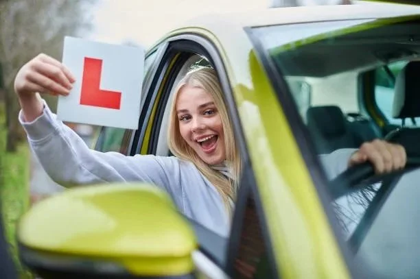 A smiling young woman holding a learner's permit sign with a red letter L, sitting in the driver's seat of a green car, celebrating a driving milestone.