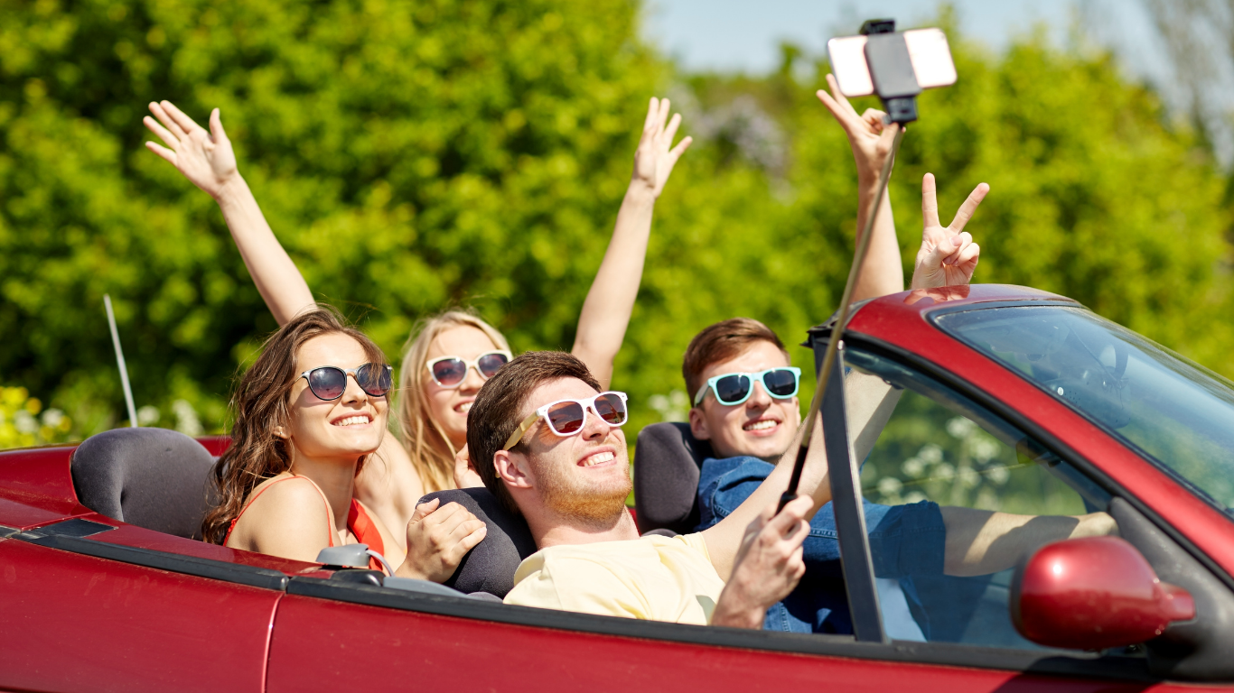 Four young friends riding in a red convertible car, taking a selfie with a smartphone, smiling, wearing sunglasses, on a sunny day with green trees in the background.