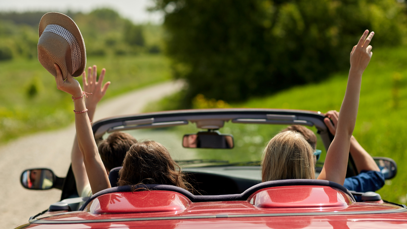 Three people are riding in a red convertible car on a countryside road, with two women raising their arms outside the car, one holding a hat, in a sunny setting with green trees and grass.
