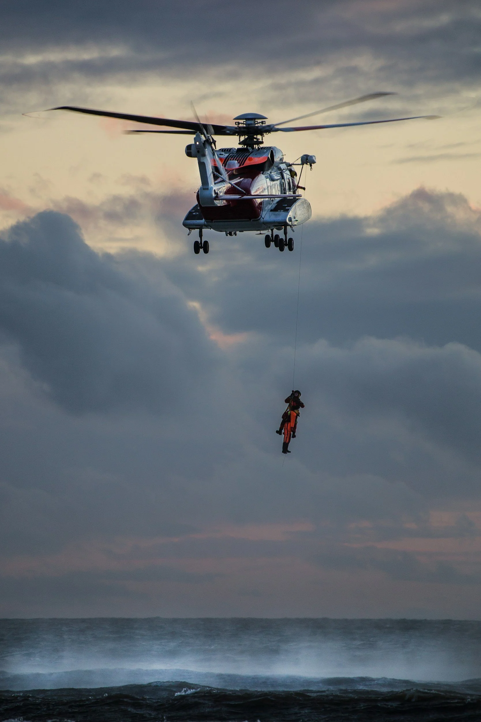 An emergency helicopter flying over the ocean with an individual dangling from a rescue line, at sunrise.