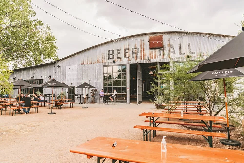 Outdoor dining area outside a rustic, metal building with a sign that says BEEHALL, featuring picnic tables, umbrellas, and a few people sitting and standing near the entrance.