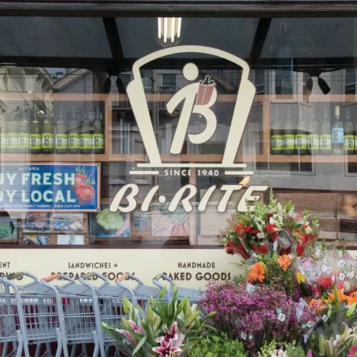 Store window display with a large BI-RITE logo, flowers, shopping carts, and signage for fresh, local, handmade baked goods.
