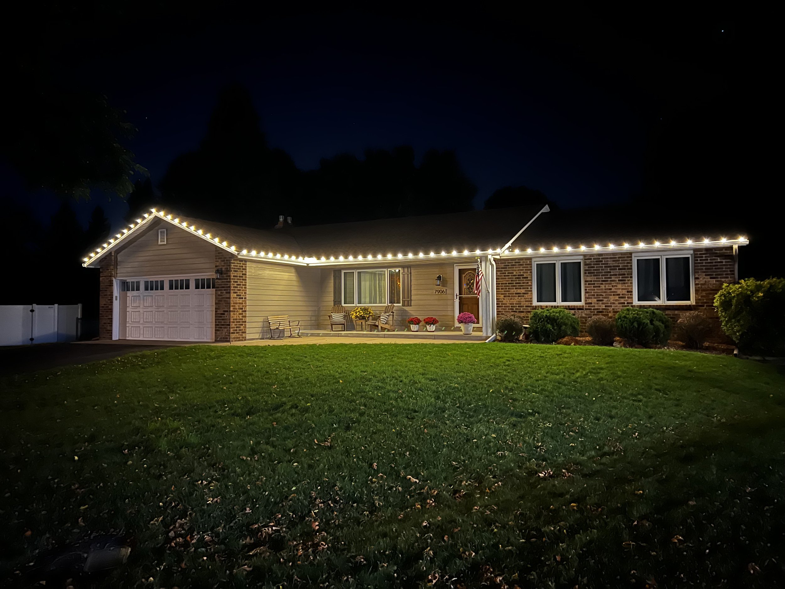 Single-story house at night decorated with white string lights along the roofline, with a well-kept lawn and outdoor seating area on the porch. The house has a brick and siding exterior with a garage, windows, and an American flag near the front door