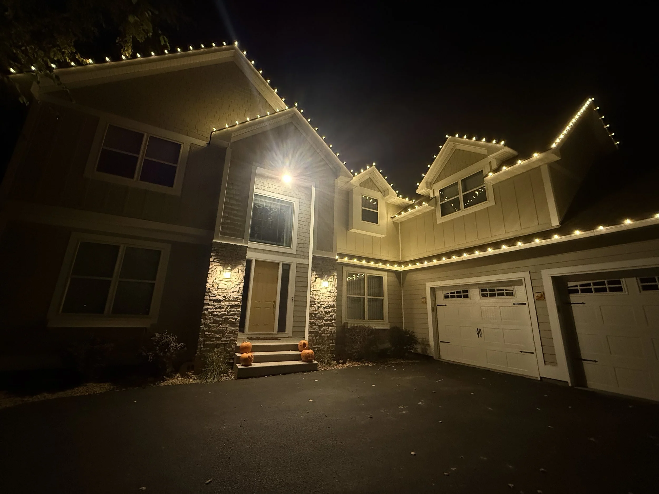 Night view of a two-story house decorated with string lights along the roofline, front steps with pumpkin decorations, and exterior lights illuminating the entrance.
