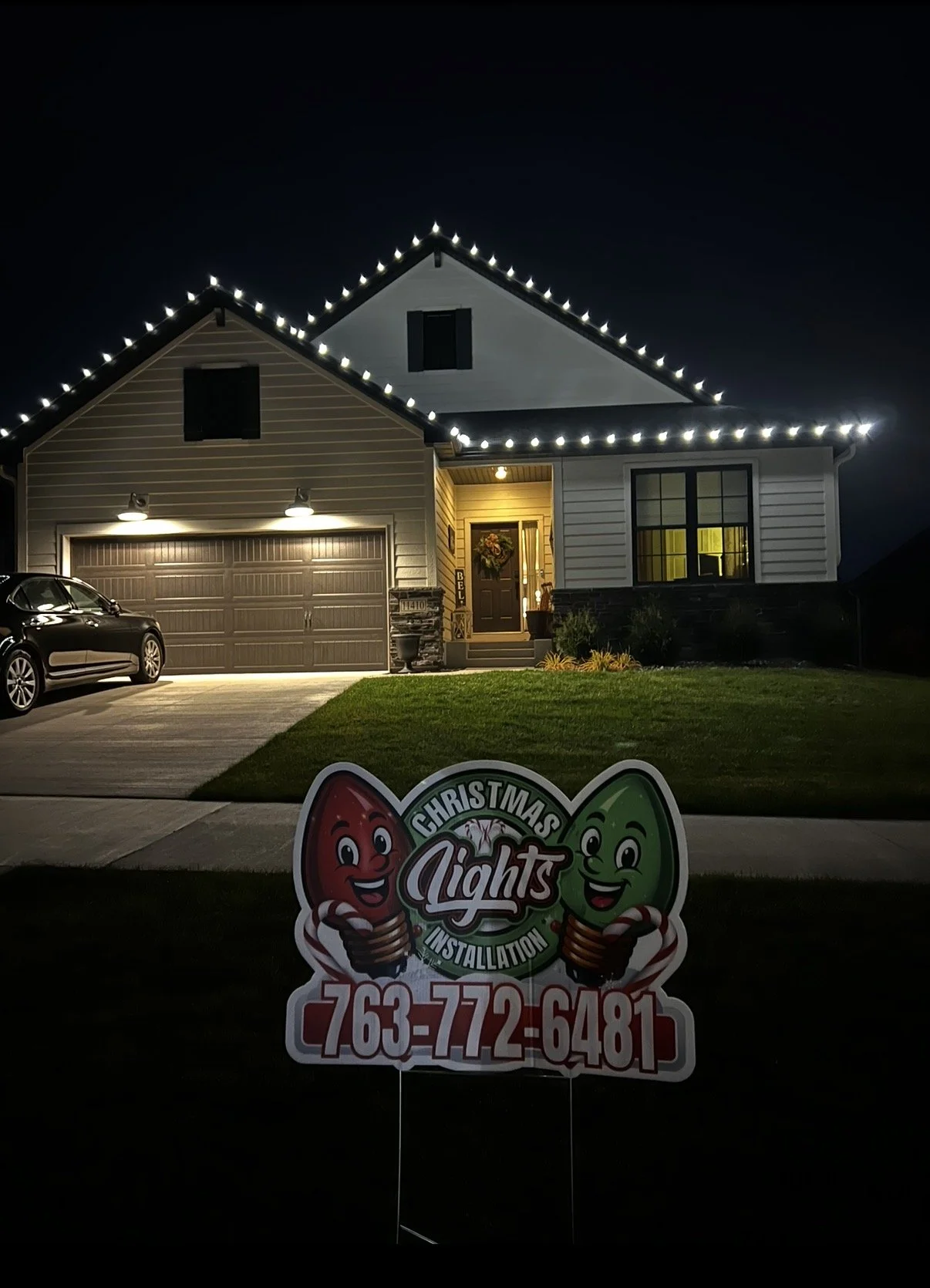 A house decorated with Christmas lights is shown at night, with a sign in front advertising Christmas light installation services and a phone number.
