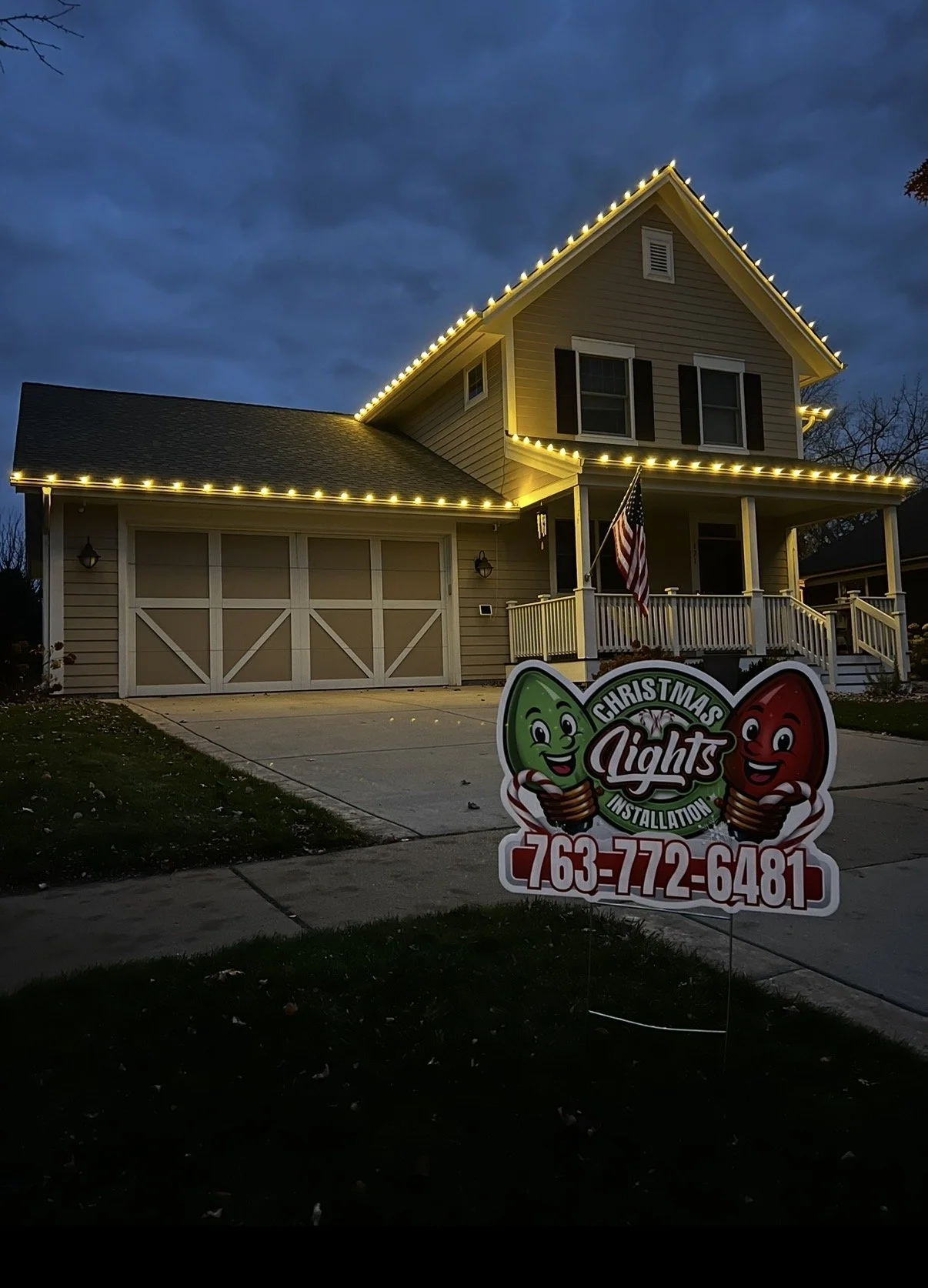 A house decorated with Christmas lights at night, with a yard sign displaying a Christmas lights installation company's contact information.