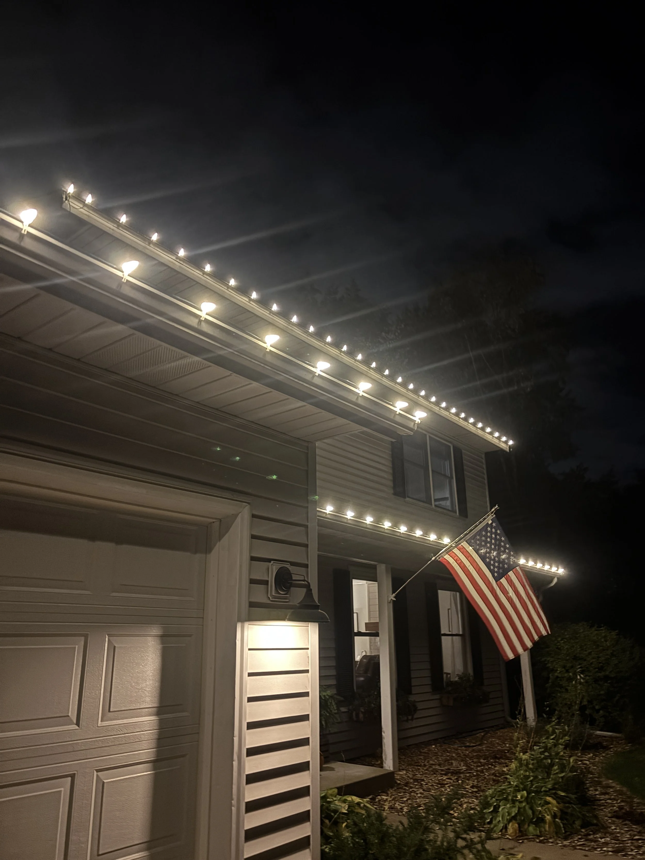 A two-story house at night with white exterior siding, illuminated by outdoor lighting, decorated with a string of lights along the roofline, and an American flag on a flagpole near the front entrance.