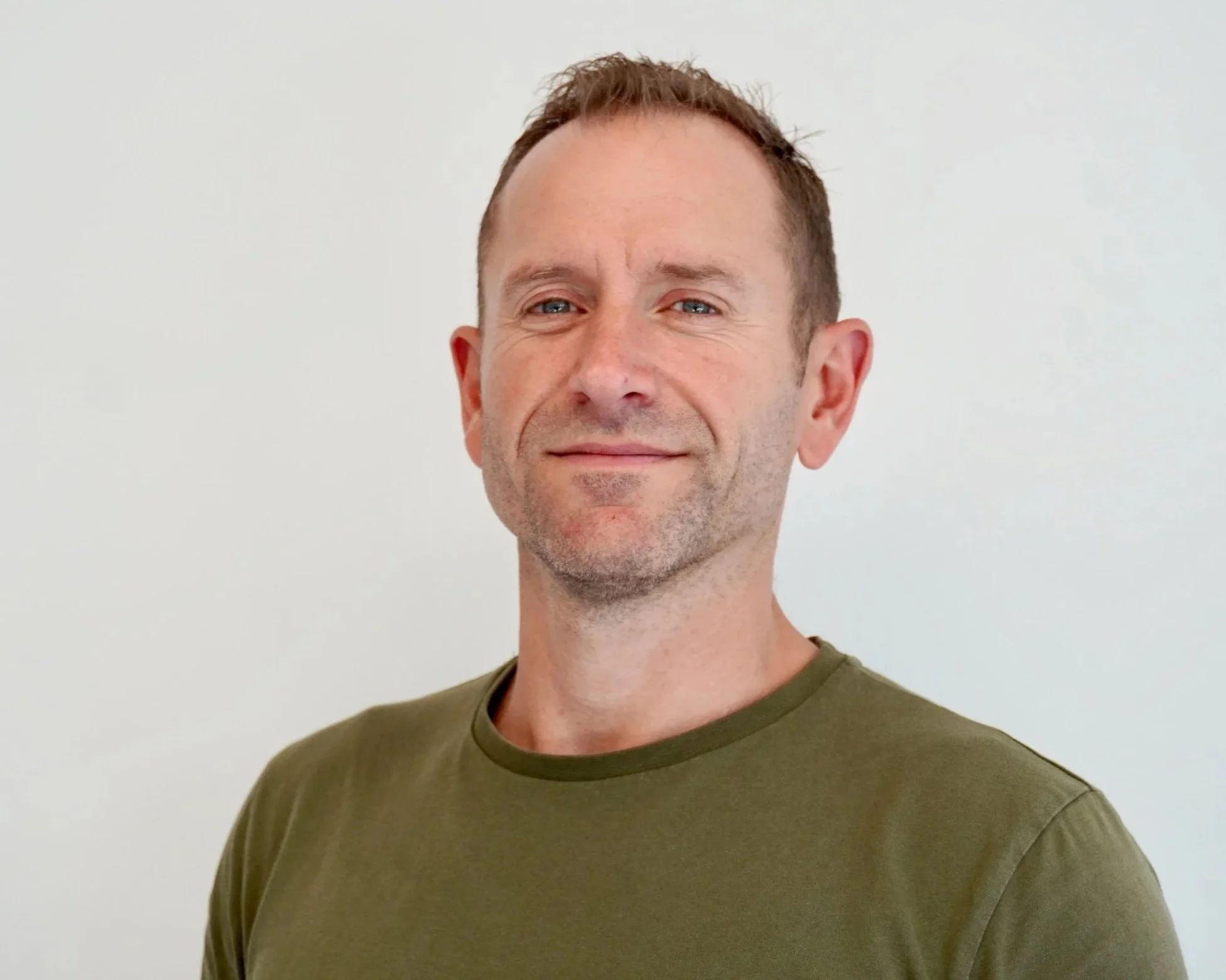 A man with short brown hair, blue eyes, and light stubble, smiling slightly and wearing a green t-shirt, standing against a plain white wall.
