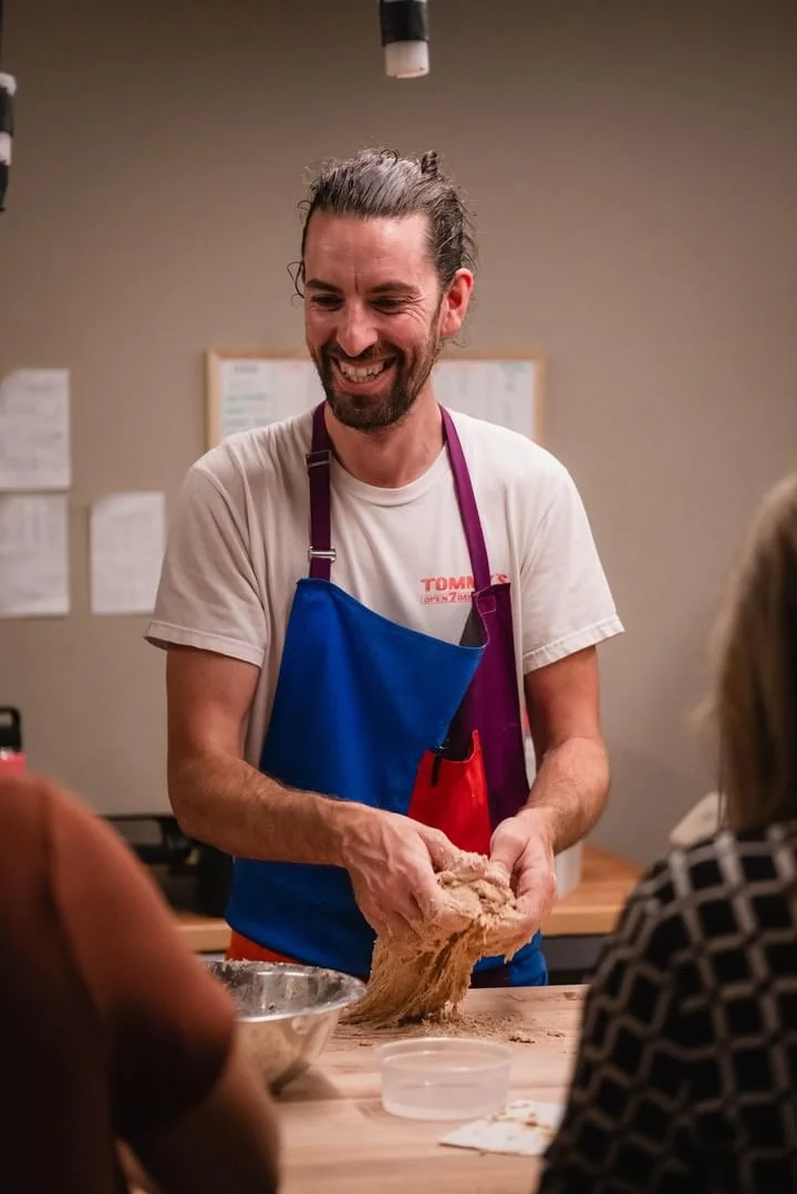 A man with long gray hair, a beard, and a smile, wearing a blue apron over a white T-shirt, is kneading dough in a kitchen or bakery setting. Two women are seated at the table, watching him.
