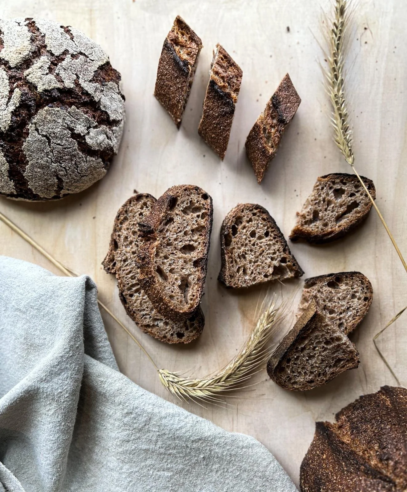 Sliced and whole rustic bread on a light wooden surface with wheat stalks and a light gray cloth.
