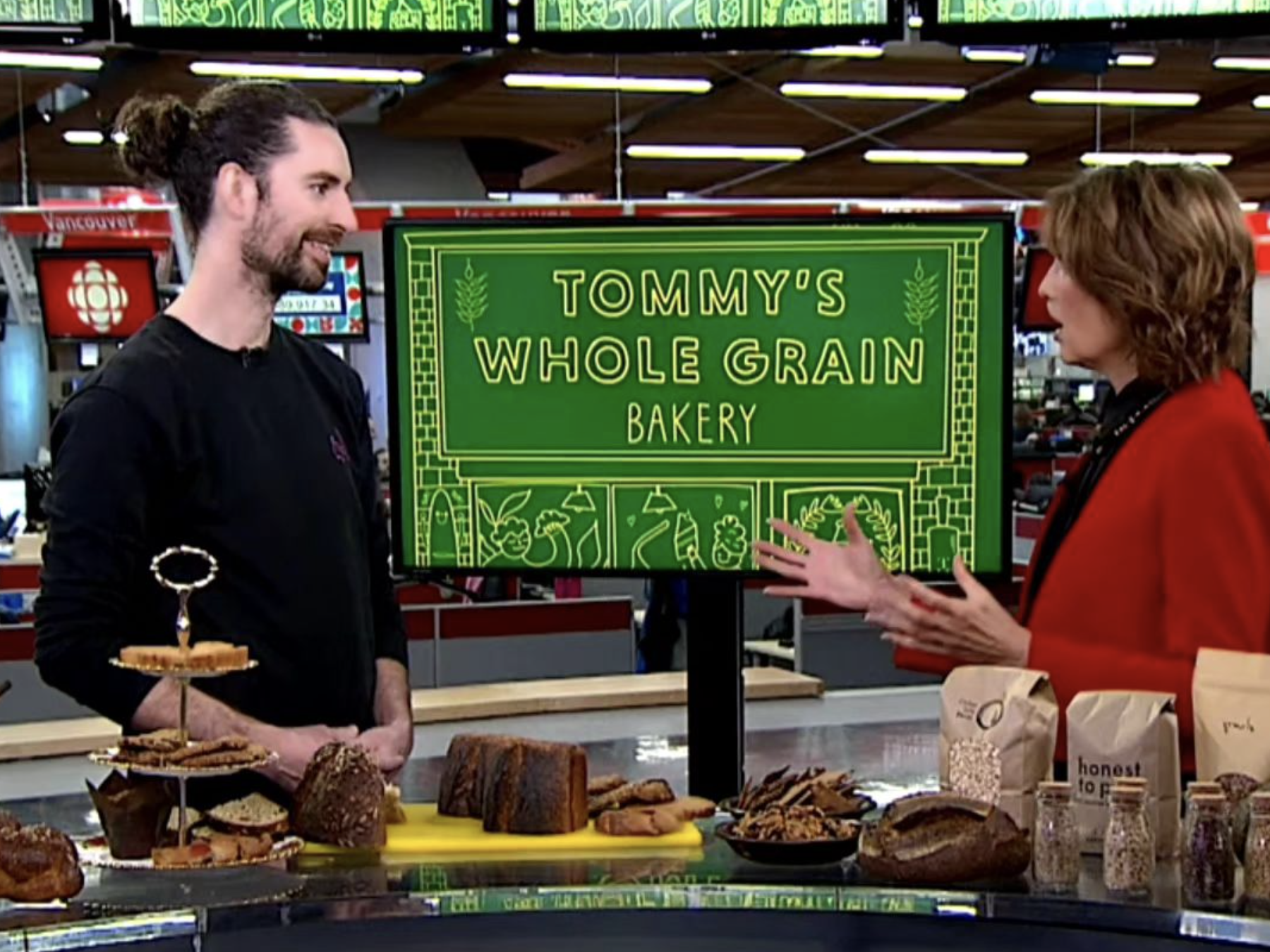 A man and a woman are talking in a bakery, with a sign that reads 'Tommy's Whole Grain Bakery' in the background. The table in front has various baked goods and grains.