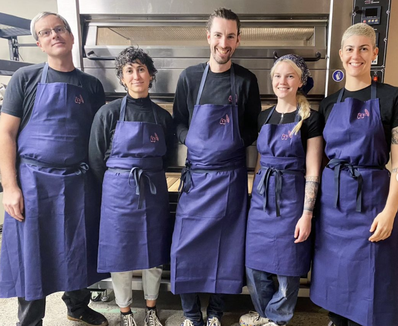 Group of five people in black shirts and blue aprons standing in a kitchen, smiling at the camera.