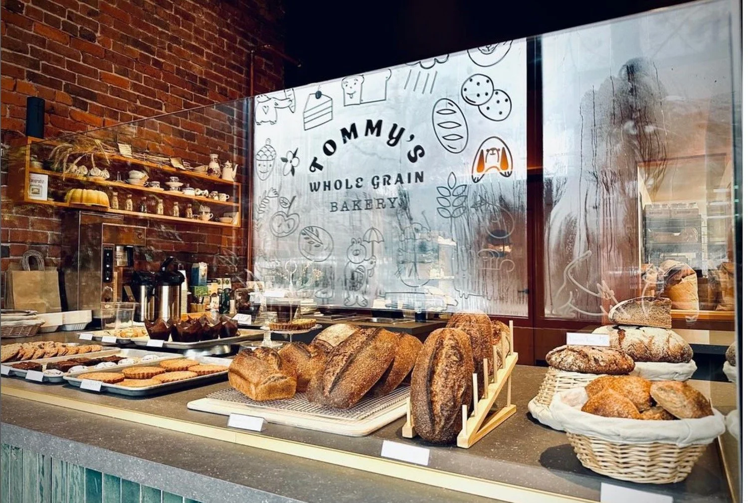 Display of freshly baked bread and pastries at Tommy's Whole Grain Bakery, with a brick wall and bakery sign in the background.