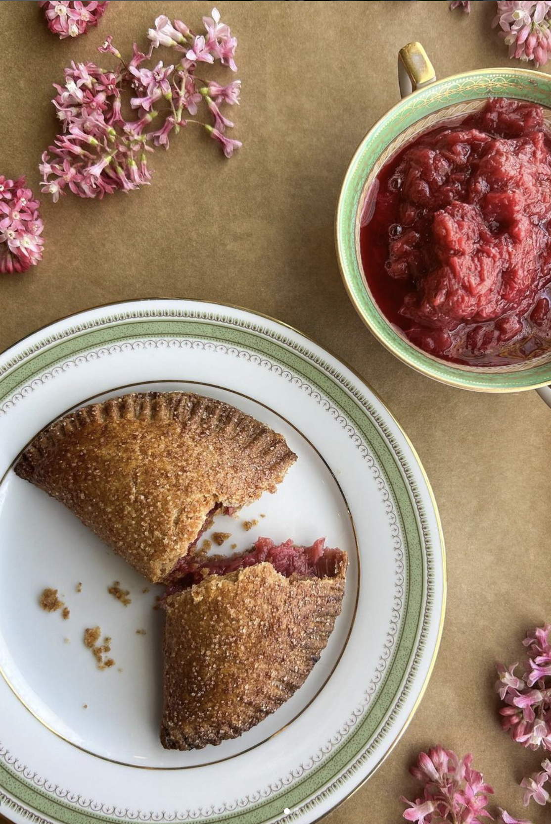 A partially eaten fruit pastry on a decorative plate, with a cup of red fruit jam or preserves and pink flowers on a brown paper background.
