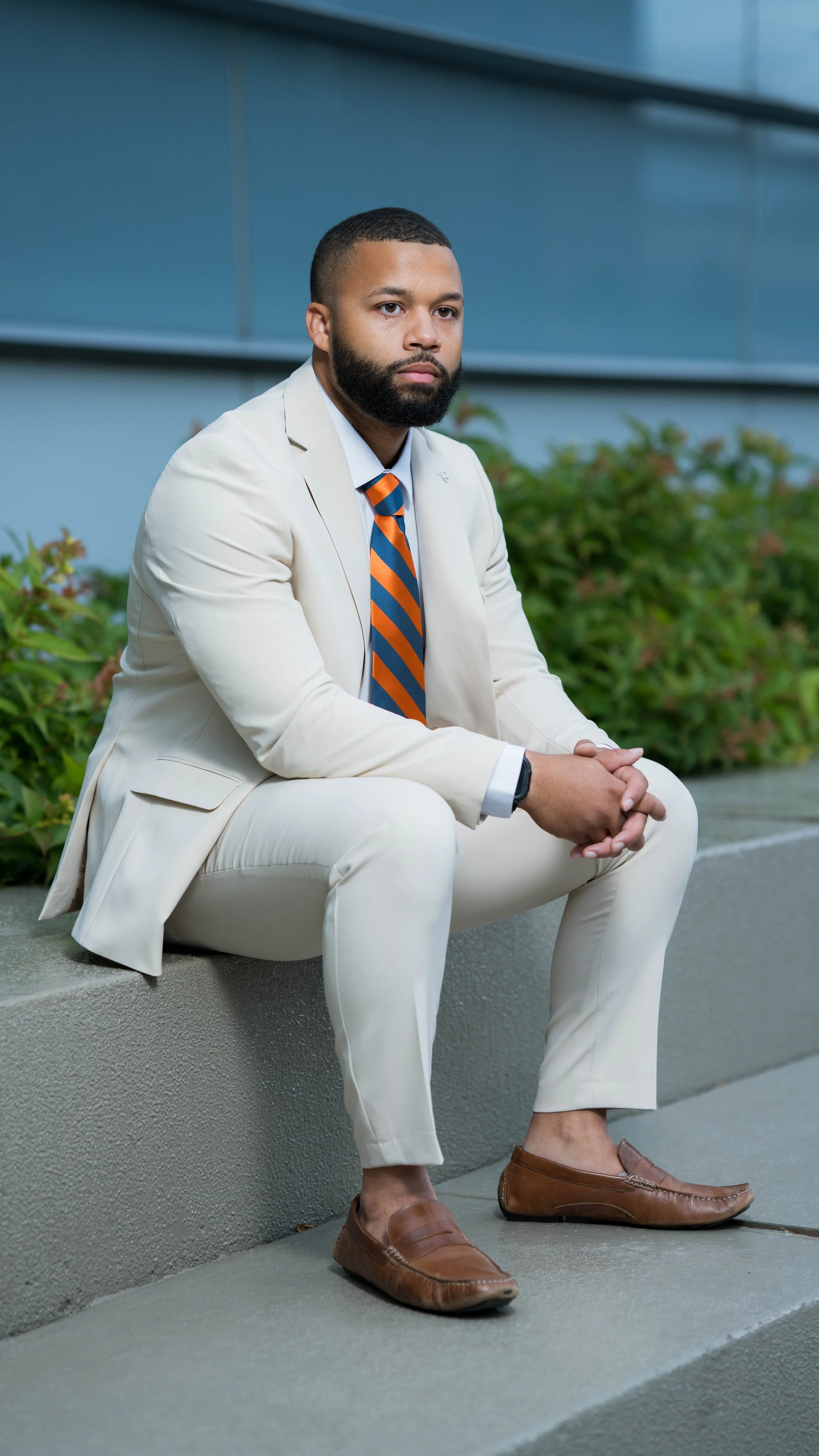 A man with a beard in a cream suit, white shirt, and striped orange and blue tie, sitting outdoors on a concrete ledge with green bushes behind him. He has short hair and is wearing brown loafers.