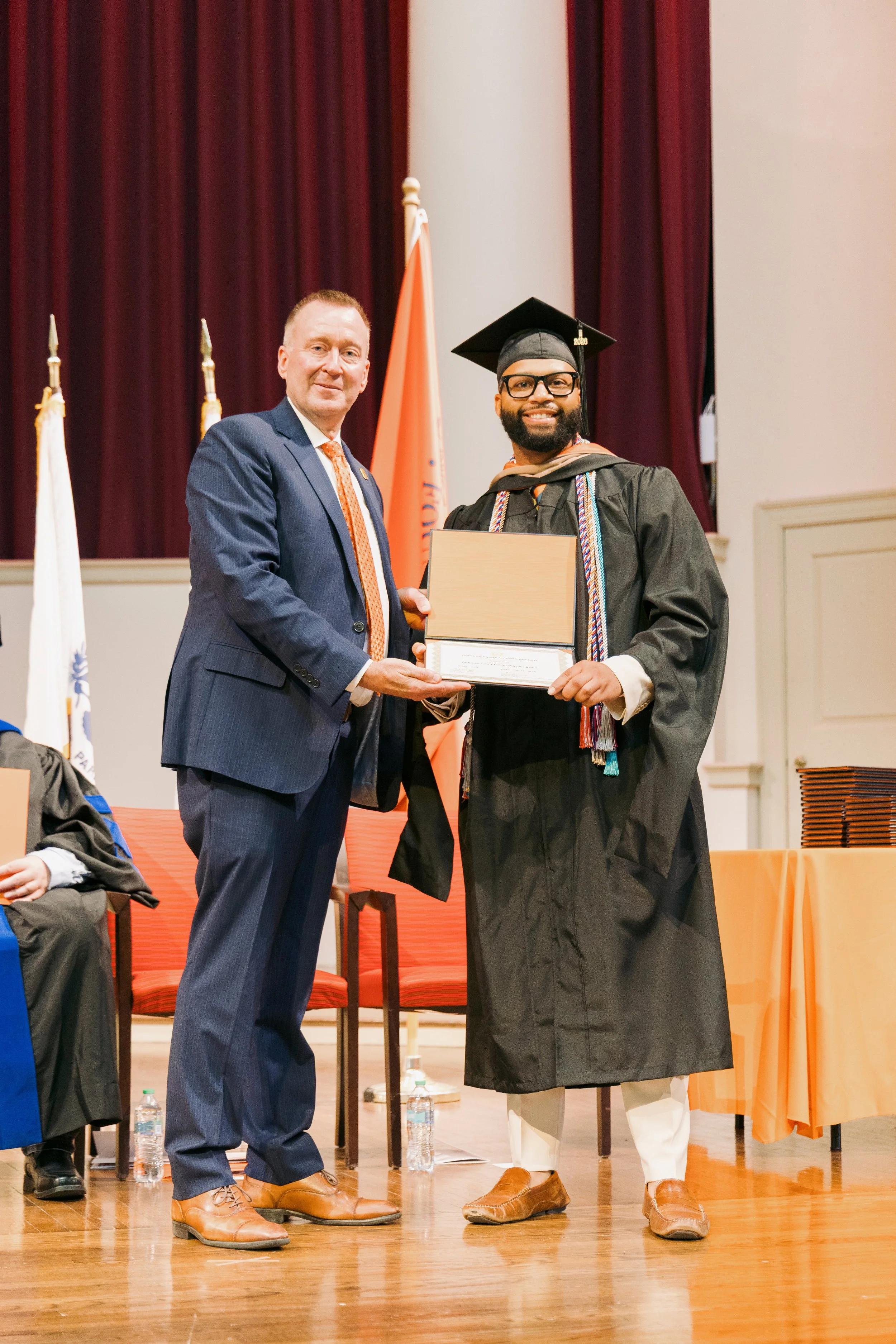 A man in a graduation cap and gown receiving a diploma from another man in a suit on stage at a graduation ceremony.