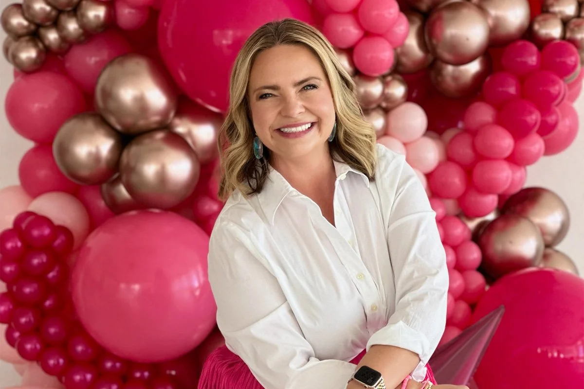 A woman with shoulder-length blonde hair smiling while standing in front of a pink and metallic balloon backdrop.